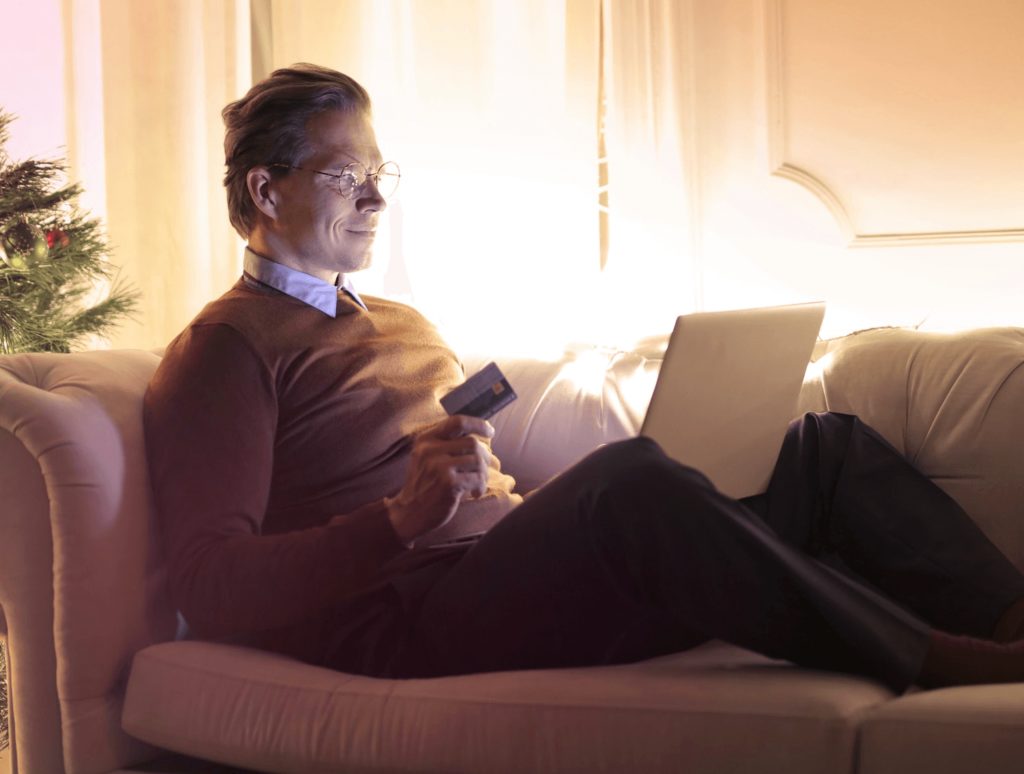 Image of a man sitting on a couch holding a credit card while shopping on a laptop